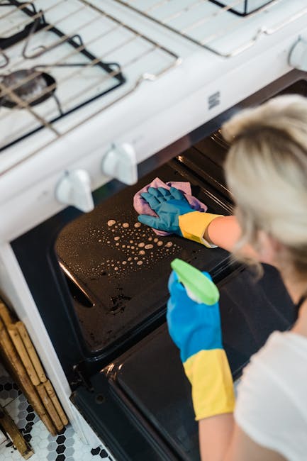 A person wearing blue and yellow rubber gloves is using a pink cleaning cloth to wipe the black oven interior, which has some visible soap or cleaning solution bubbles on the surface. The oven door is open, revealing a clean, reflective metal interior. The surrounding area includes a white gas stove with black grates and control knobs, and a portion of a tiled floor with a black and white pattern is visible. The lighting is bright, highlighting the shiny surface of the oven and the overall cleanliness. This image reflects professional surface cleaning and sanitisation processes, emphasizing thorough deep cleaning of kitchen appliances. The portrayal aligns with the services offered by Cleaners Kensington for residential or commercial oven cleaning, fitting naturally with the page on affordable oven cleaning quotes for Kensington businesses.