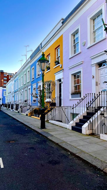 A row of colorful Georgian-style terraced houses on a residential street, painted in shades of blue, yellow, and lavender, with white-framed windows and black wrought-iron railings along the steps leading to front doors. The pavement is clean, and a traditional black lamppost is positioned near the curb, under a bright blue sky. The image reflects a vibrant neighborhood, with neat outdoor areas, perfect for professional domestic cleaning or surface cleaning services promoted by Cleaners Kensington, as mentioned on the page titled 'Affordable oven cleaning quotes for Kensington businesses, Kensington'.