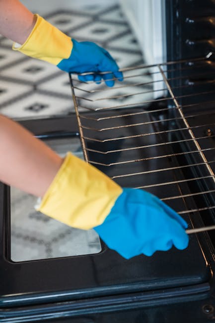 A person wearing blue and yellow rubber gloves is using a pink cleaning cloth to wipe the black oven interior, which has some visible soap or cleaning solution bubbles on the surface. The oven door is open, revealing a clean, reflective metal interior. The surrounding area includes a white gas stove with black grates and control knobs, and a portion of a tiled floor with a black and white pattern is visible. The lighting is bright, highlighting the shiny surface of the oven and the overall cleanliness. This image reflects professional surface cleaning and sanitisation processes, emphasizing thorough deep cleaning of kitchen appliances. The portrayal aligns with the services offered by Cleaners Kensington for residential or commercial oven cleaning, fitting naturally with the page on affordable oven cleaning quotes for Kensington businesses.