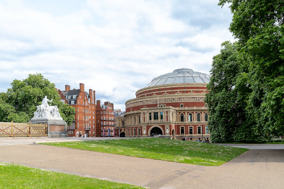 Exterior view of a large, historic concert hall with a rounded brick façade and arched windows, situated in a park with green grass and trees on either side. The building features a prominent domed r