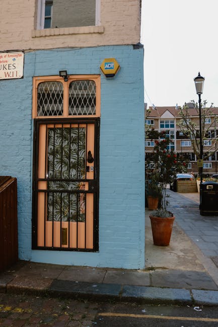 This image shows the exterior of a residential building on Gloucester Road, featuring a blue-painted brick wall with a black metal security door that has decorative iron bars and transparent glass panels with a leafy pattern. Above the door, there are two small, arched windows with decorative metal grilles. To the right side of the building, outdoor pavement extends into a street with parked cars, a streetlamp, and potted plants, including a large terracotta planter with a shrub. The scene is lit by natural daylight, highlighting the clean, well-maintained surfaces and the fresh appearance of the building's facade, suggesting a recent deep cleaning or maintenance. For a thorough surface cleaning and sanitisation of similar residential exteriors, contact Cleaners Kensington, experts in domestic cleaning services.