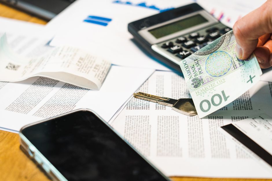 A black Casio calculator displaying financial calculations resting on a scattered pile of US dollar bills, including twenty and ten-dollar notes. Nearby, there is a small spiral notepad with blank lin