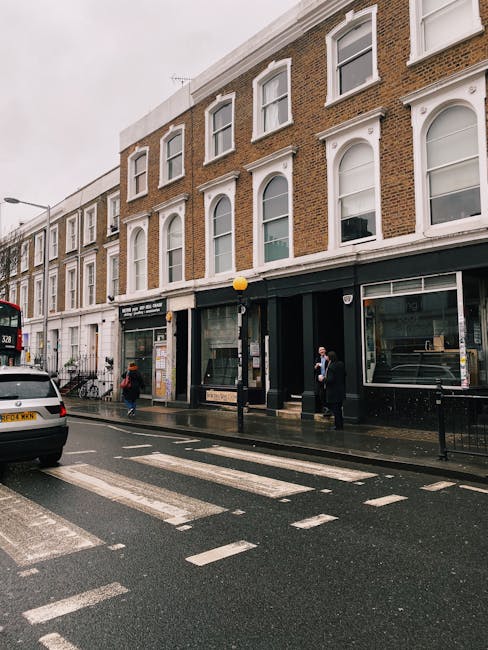 A street view of a multi-story building on Kensington High Street featuring a ground-level commercial space with large glass windows and black framing. The building's facade is constructed of red brick with white decorative window frames on the upper floors. The sidewalk in front is wet, indicating recent cleaning or rain, and there are a few pedestrians walking and standing near the shopfronts, some wearing masks. A white van and a bus are visible on the street, and a zebra crossing is in the foreground. The scene is captured under overcast sky lighting, emphasizing the clean and well-maintained appearance of the area, consistent with professional deep cleaning and surface sanitation practices provided by Cleaners Kensington for maintaining hygiene in commercial properties.