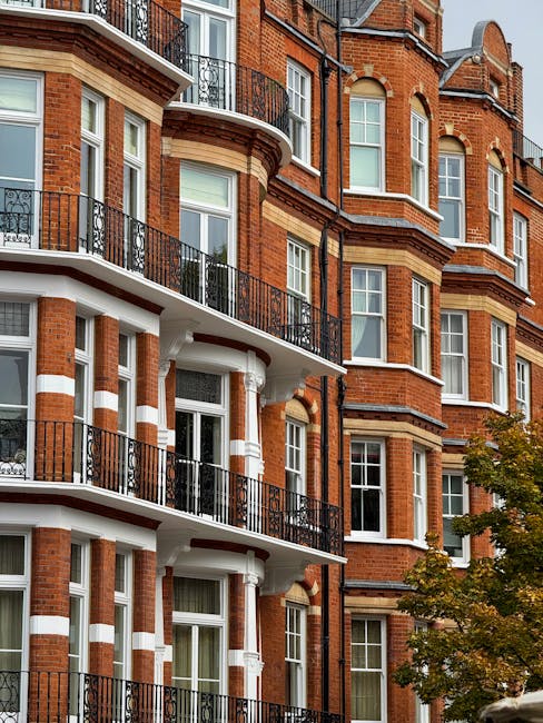 Exterior view of a multi-story red brick residential building with white decorative accents and black iron balcony railings. The building features large, evenly spaced white-framed sash windows, some with visible curtains or blinds. The balconies are lined with clean, polished black metal railings, and the surface of the balconies appears well-maintained and free of debris. The lighting suggests daylight, highlighting the building's architectural details and the lush green foliage of nearby trees. This image exemplifies the importance of regular surface cleaning and maintenance in residential properties, as promoted by Cleaners Kensington, to ensure a tidy and hygienic environment.