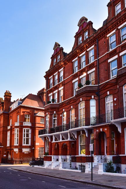 A street view of a multi-story building on Kensington High Street featuring a ground-level commercial space with large glass windows and black framing. The building's facade is constructed of red brick with white decorative window frames on the upper floors. The sidewalk in front is wet, indicating recent cleaning or rain, and there are a few pedestrians walking and standing near the shopfronts, some wearing masks. A white van and a bus are visible on the street, and a zebra crossing is in the foreground. The scene is captured under overcast sky lighting, emphasizing the clean and well-maintained appearance of the area, consistent with professional deep cleaning and surface sanitation practices provided by Cleaners Kensington for maintaining hygiene in commercial properties.