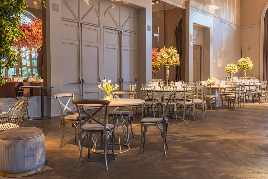 An elegantly set event space with round tables covered in white tablecloths, each adorned with tall vases of pink and white floral arrangements. The room features polished wooden flooring and cream-colored walls with decorative paneling. Natural light filters through large windows, casting a warm glow over the interior. Multiple chairs with metal frames and cushioned seats are arranged around the tables, and in the foreground, there is a small round table with a yellow flower centerpiece and a nearby upholstered ottoman. On the side, a sideboard or serving area with glassware and candles is visible. The space appears clean and well-maintained, reflecting the professional surface cleaning, deep cleaning, and sanitisation services provided by Cleaners Kensington for the Kensington Palace event cleanup case study.