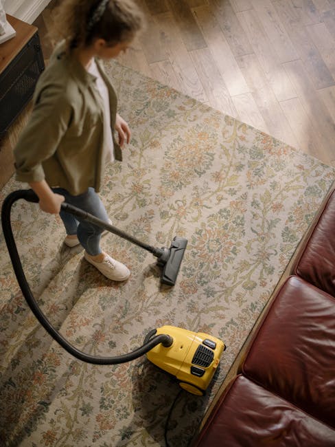 A woman using a yellow vacuum cleaner on a patterned area rug with floral motifs in a living room. The floor underneath appears to be wooden or laminate, with a brown leather sofa visible on the right side. The woman is wearing a beige jacket, light-colored jeans, and white sneakers, and is focused on cleaning the rug. The room is well-lit, highlighting the cleanliness and freshness of the space, which is being maintained by Cleaners Kensington through surface cleaning and deep cleaning processes to ensure hygiene and a tidy environment.