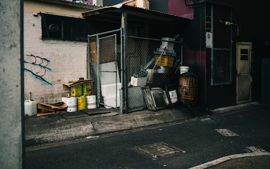 An outdoor area adjacent to a building features a storage shed made of metal with a chain-link door, containing various cleaning supplies, tools, and containers. Outside the shed, there are several yellow and white paint buckets, some cardboard boxes, and a white plastic container. The ground is paved with asphalt, with a curb separating the paved surface from the shed. The building's exterior wall is painted white with some graffiti art, and part of a window with a black security grille is visible above the shed. The scene is lit by natural daylight, highlighting the orderly yet utilitarian setup, which suggests a focus on maintenance and cleanliness as part of property management. For residents or property managers in Kensington, Cleaners Kensington emphasizes the importance of proper waste disposal and surface cleaning to adhere to council regulations, especially regarding bulky waste disposal, ensuring hygienic and well-maintained communal spaces.