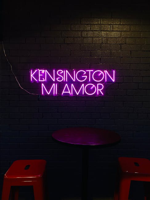 A dark interior wall featuring a bright purple neon sign that reads 'KENSINGTON MI AMOR', illuminating the surrounding area. Below the sign is a small, round table with a smooth, reflective surface, positioned between two red stools made of durable plastic or metal. The lighting emphasizes the contrast between the vibrant signage and the darker texture of the brick wall. This setting appears to be a modern, stylish space, possibly in a commercial or residential lounge area, with a clean and well-maintained appearance. The image reflects an ambient, trendy environment that might benefit from professional deep cleaning and surface sanitisation, as provided by Cleaners Kensington, especially to maintain cleanliness in high-traffic or public spaces.